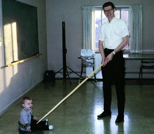 Historic photo of a missionary mopping a floor with a child riding on the mop for fun.
